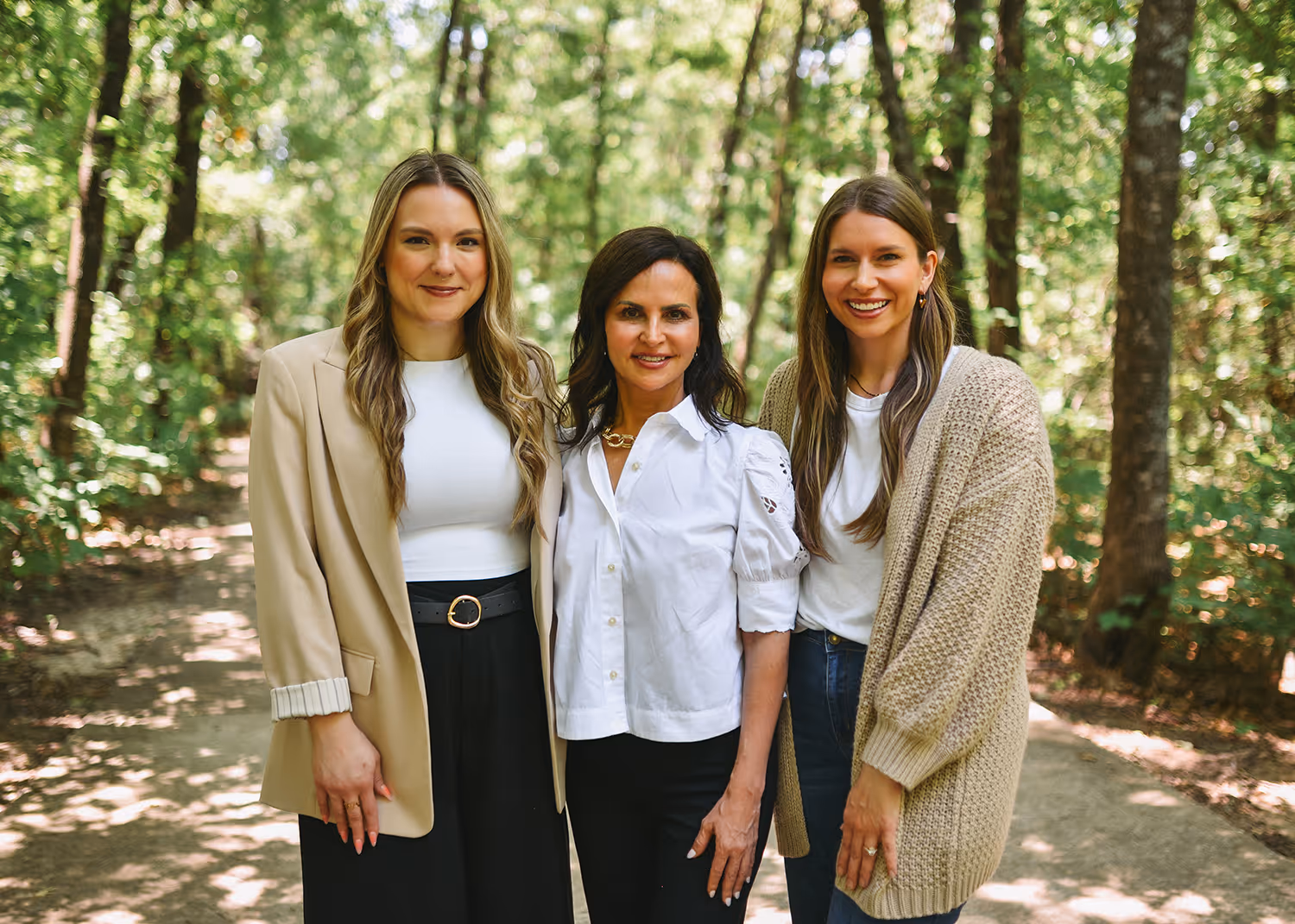 Three smiling women standing close together on a forest path with trees and sunlight in the background.