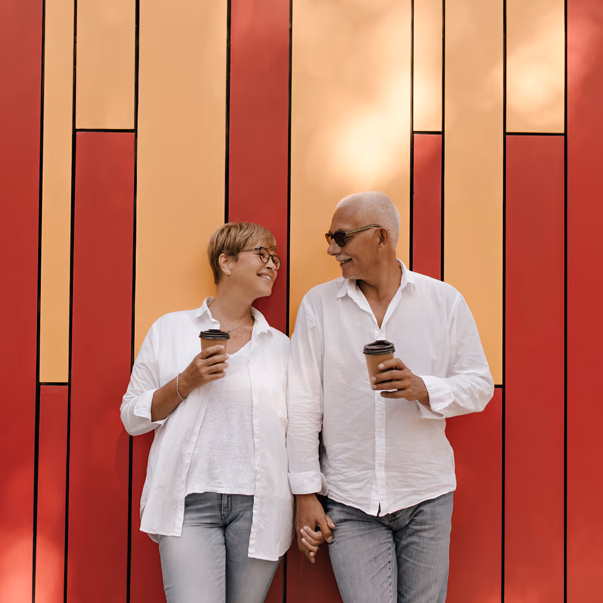 Smiling mature couple holding hands and coffee cups, standing against a red and orange wall.