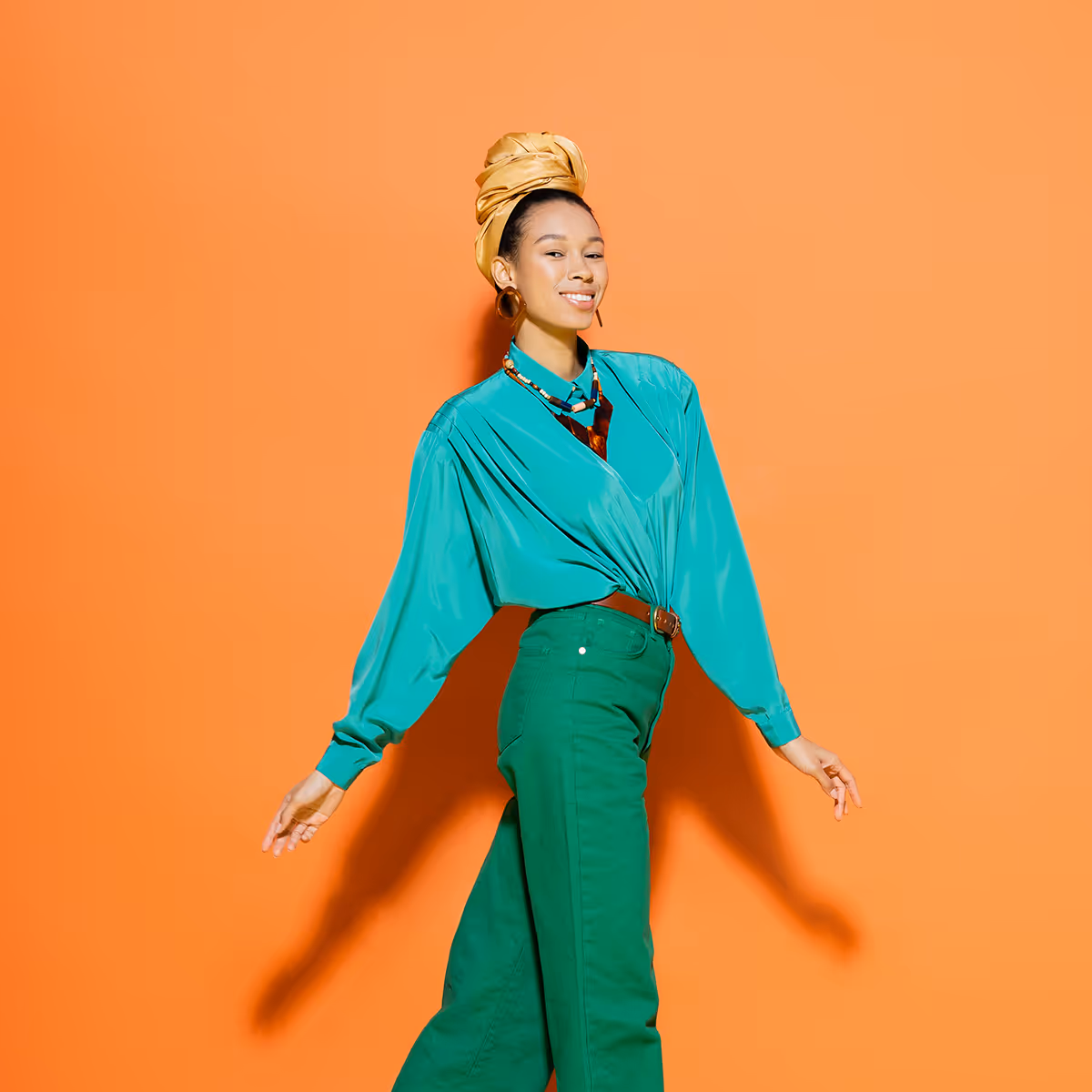 Smiling woman in teal blouse and green pants, wearing gold headwrap, posing against orange background.