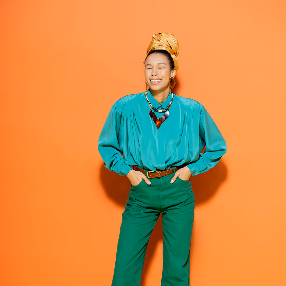 Smiling woman with eyes closed wearing turquoise shirt, green pants, gold head wrap, and necklace on orange background.