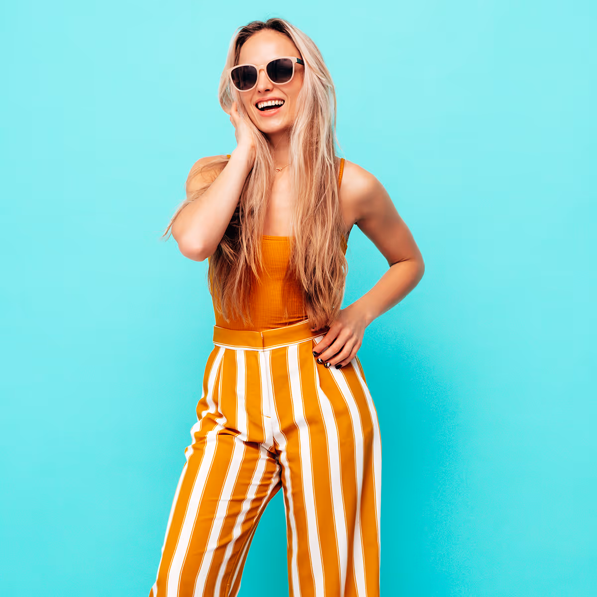 Smiling woman in sunglasses wears an orange top and striped yellow-white pants on turquoise background.
