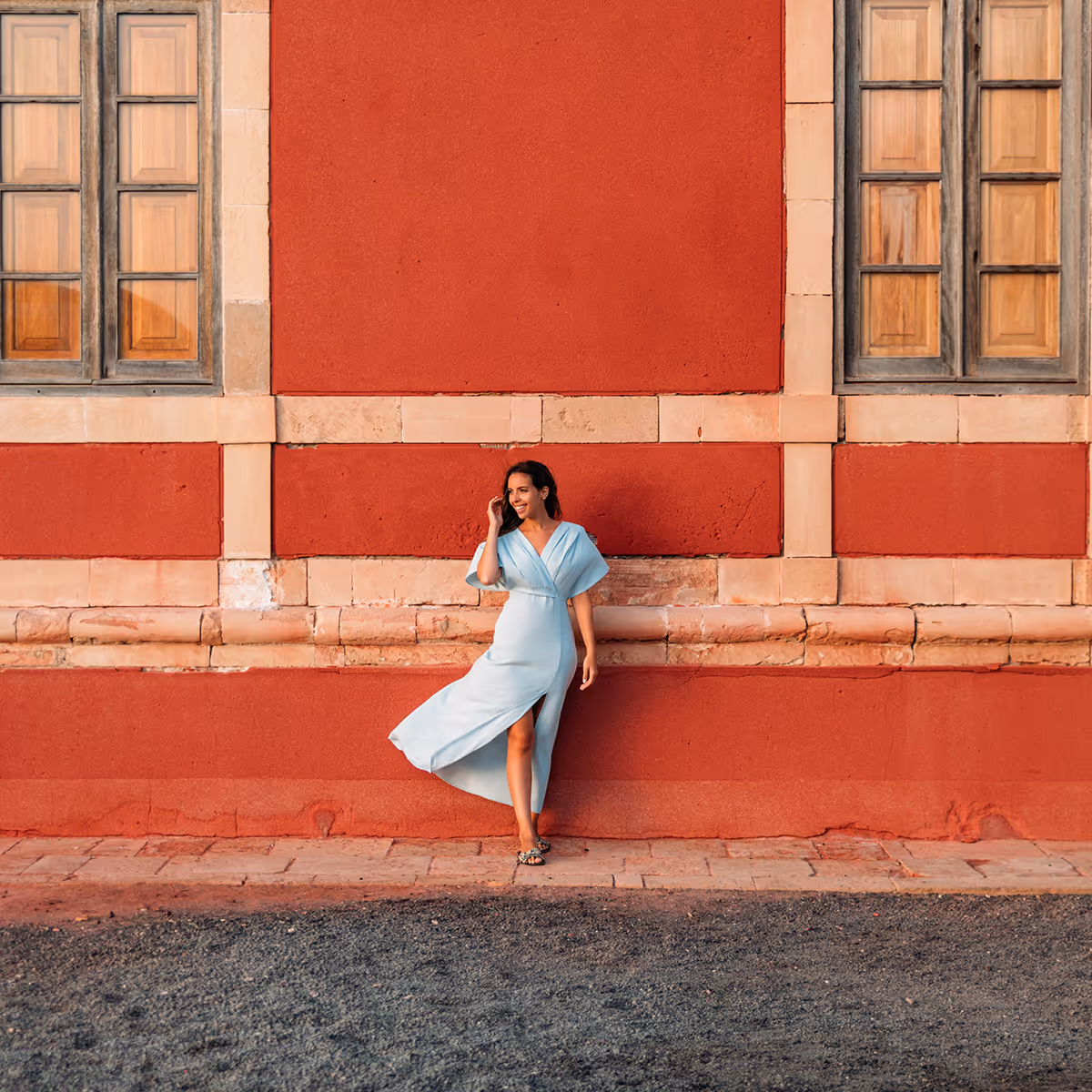 Woman in light blue dress standing and smiling against a red textured wall with windows.