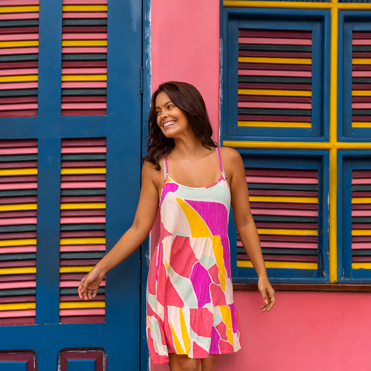 Smiling woman in colorful dress standing by a pink wall with blue, yellow, and pink shuttered windows.