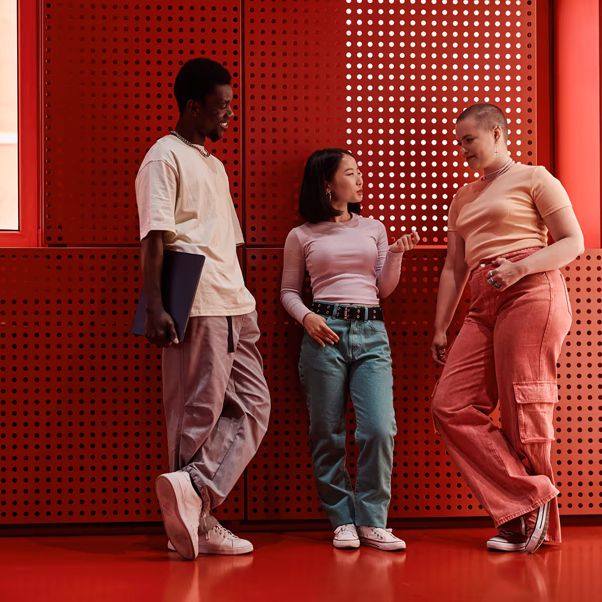 Three young adults standing and talking against a red perforated wall in casual clothing.