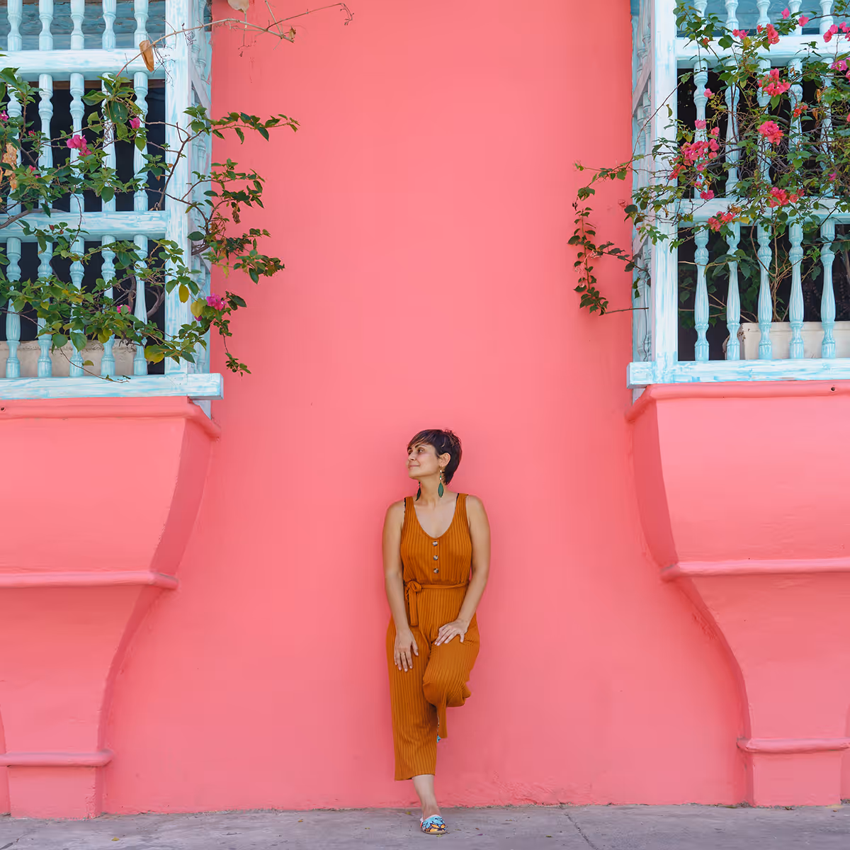 Woman in orange jumpsuit leaning against a bright pink wall between two balconies with green plants.