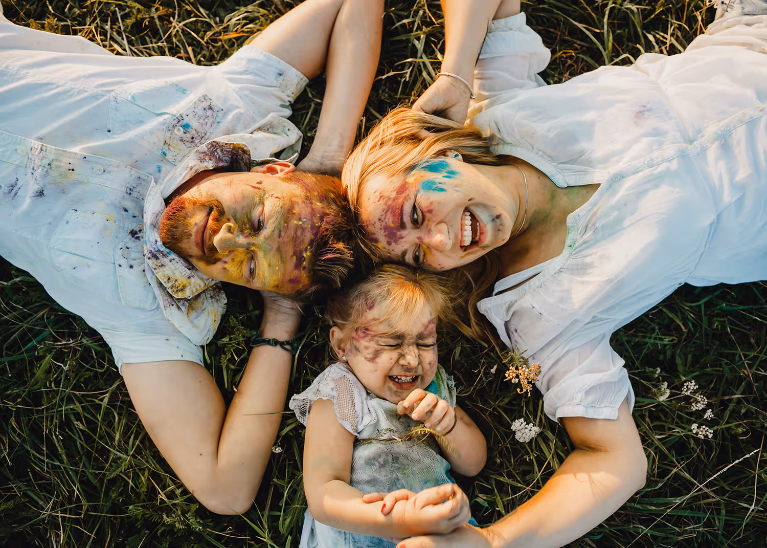 Smiling family of three with colorful paint on faces lying on grass in a playful moment.