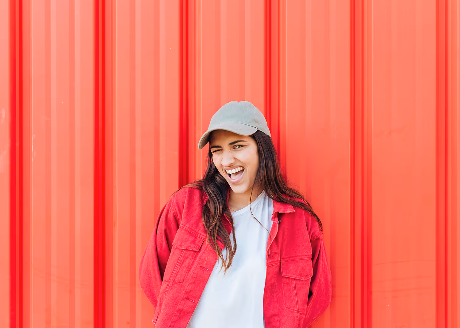 Smiling young woman winking wearing gray cap and red jacket against a red vertical panel wall.