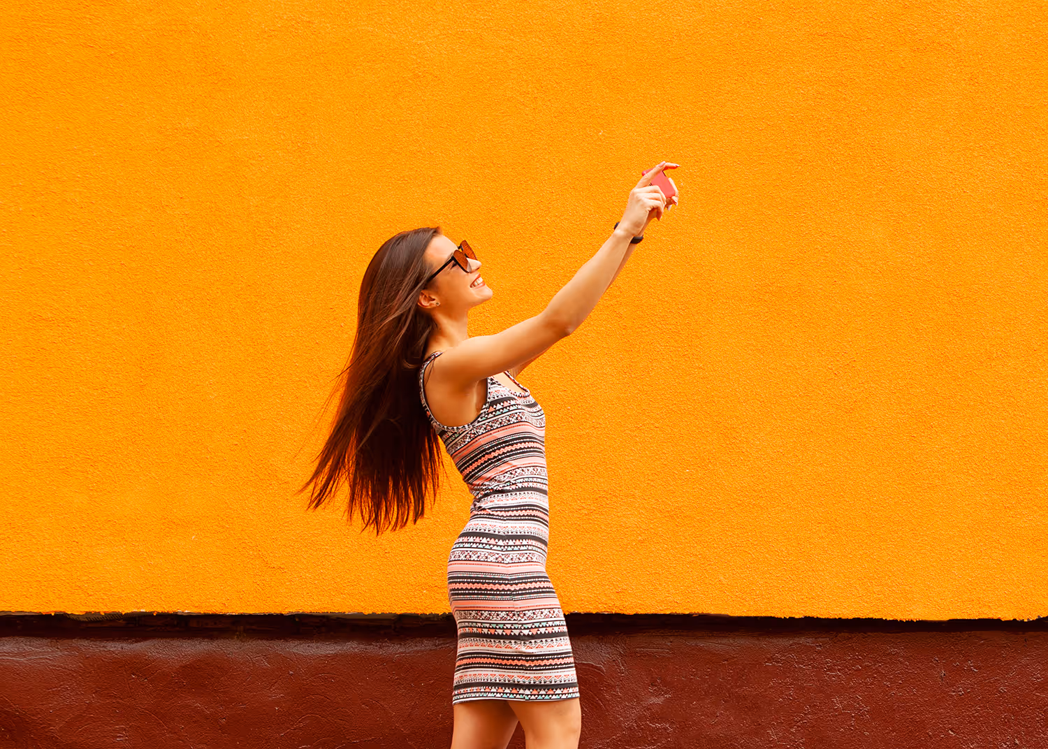 A smiling woman in a patterned dress and sunglasses takes a selfie against an orange wall.