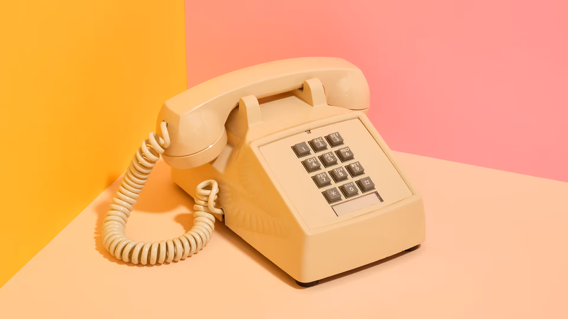 Beige vintage rotary phone with coiled cord on a pink and orange background.