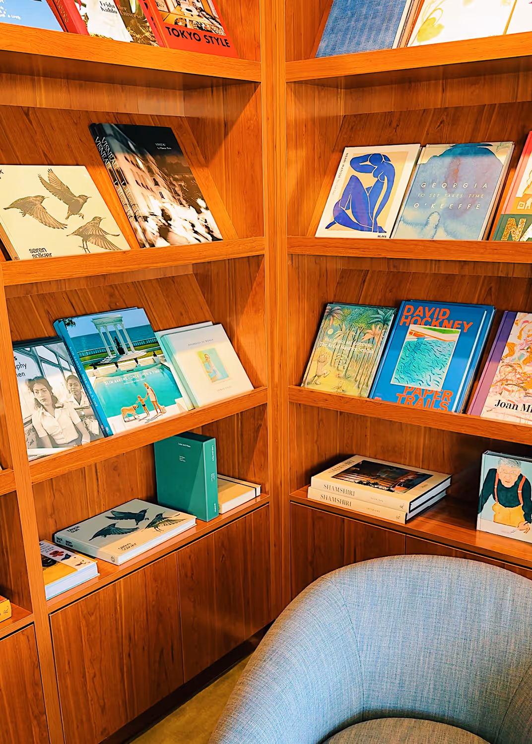 Corner wooden bookshelf filled with art and photography books, next to a gray cushioned chair.