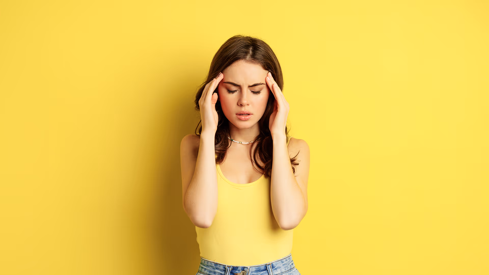 A woman holding her head in overwhelming stress or a headache against a solid yellow backdrop, illustrating the mental burden of anxiety and depression support at New Narratives Counseling.