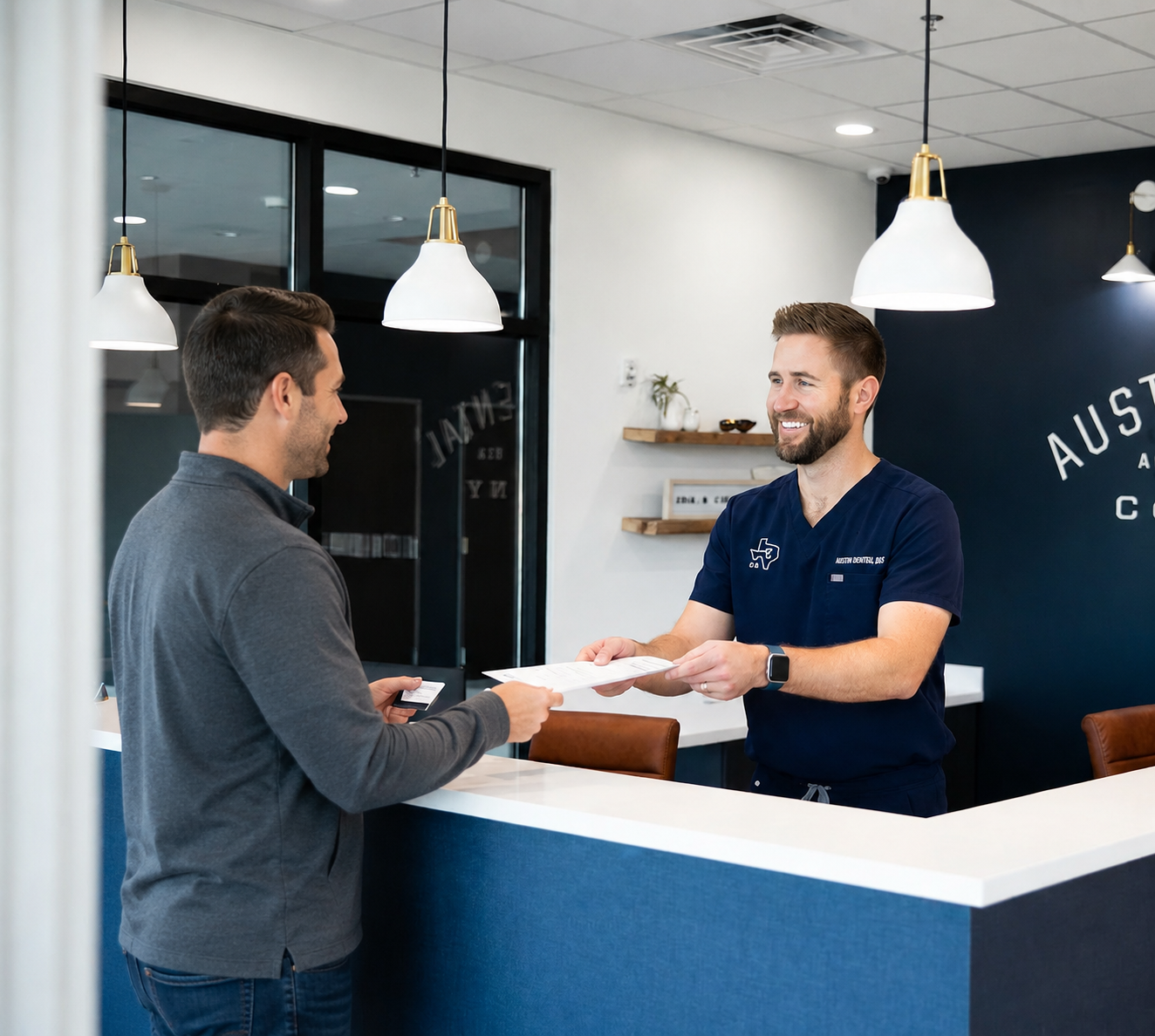 A dentist welcomes a male patient at the front desk while reviewing and exchanging appointment paperwork in a modern dental office