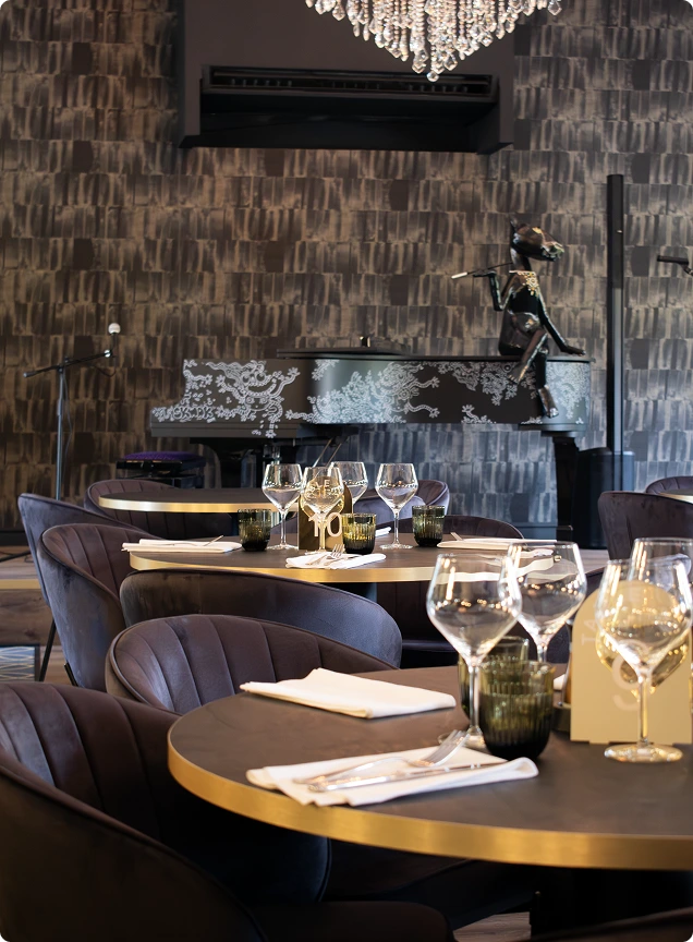 Salle du restaurant cosy, avec des tables rondes au centre et un grand piano dans le fond, sur la scene.