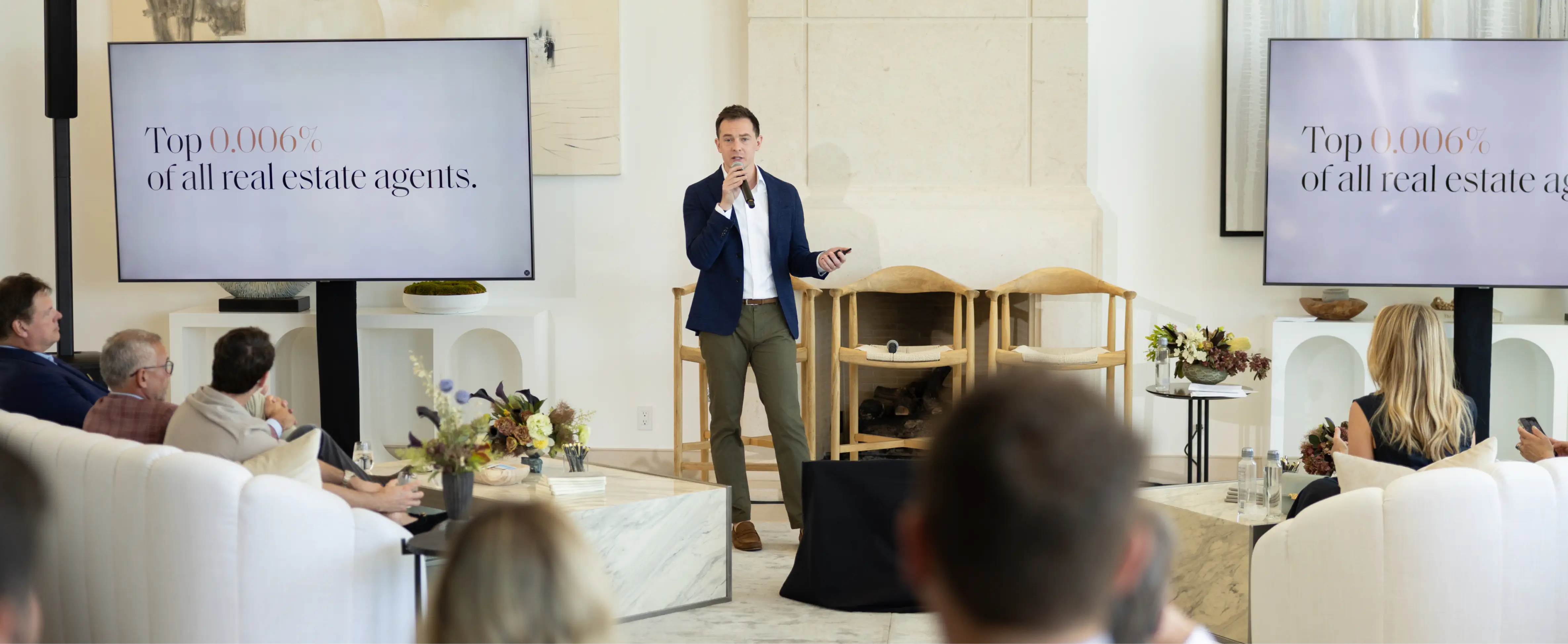 Man in blazer speaking to seated audience with two screens showing 'Top 0.006% of all real estate agents.'