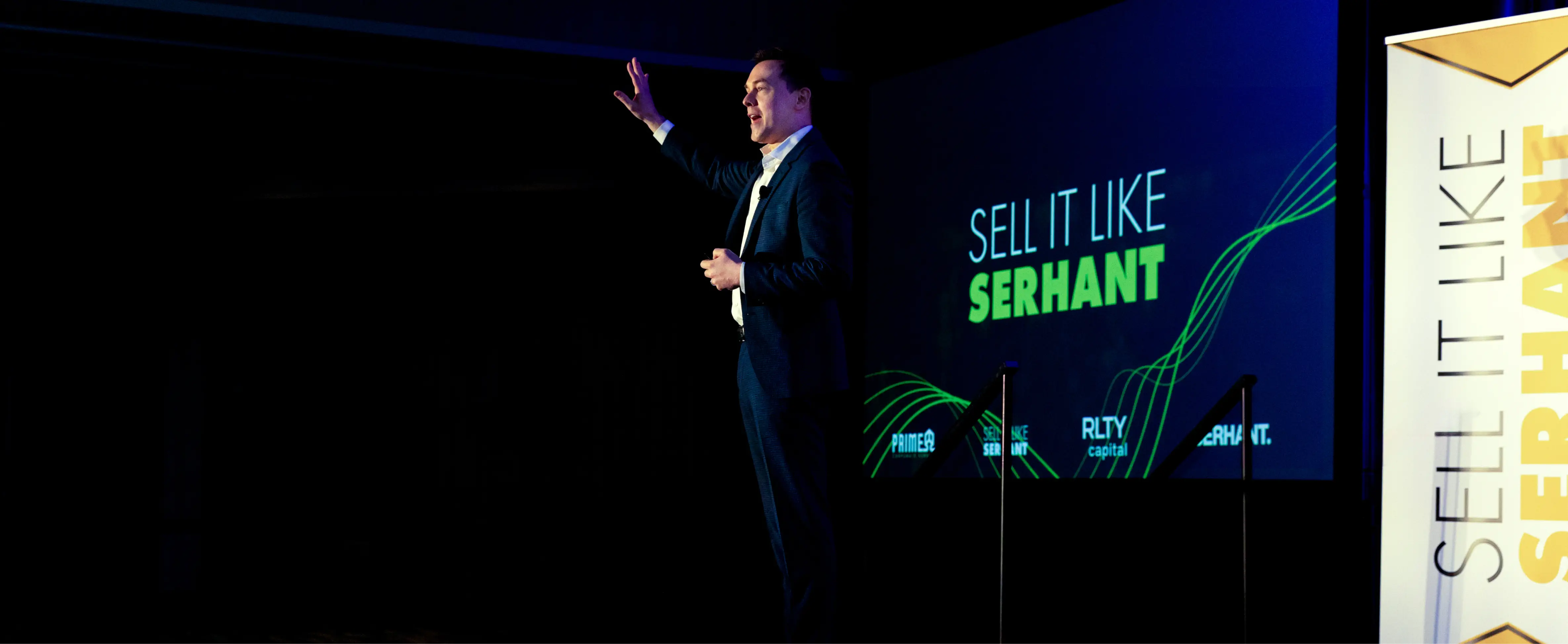 Man in suit speaking on stage with 'Sell It Like Serhant' text projected behind him.