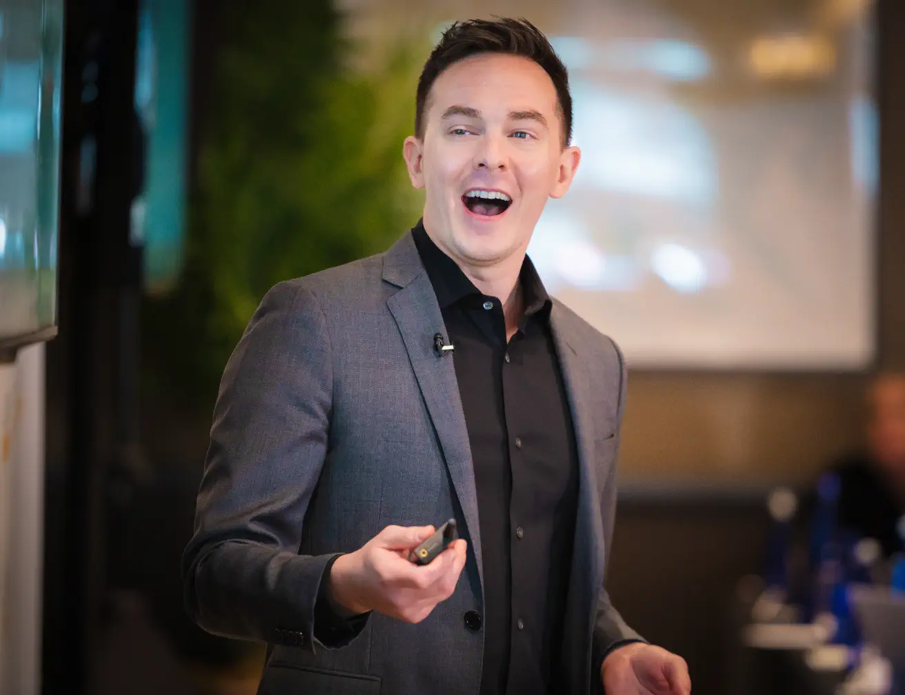Man in gray suit and black shirt speaking or presenting with a pointer in hand indoors.