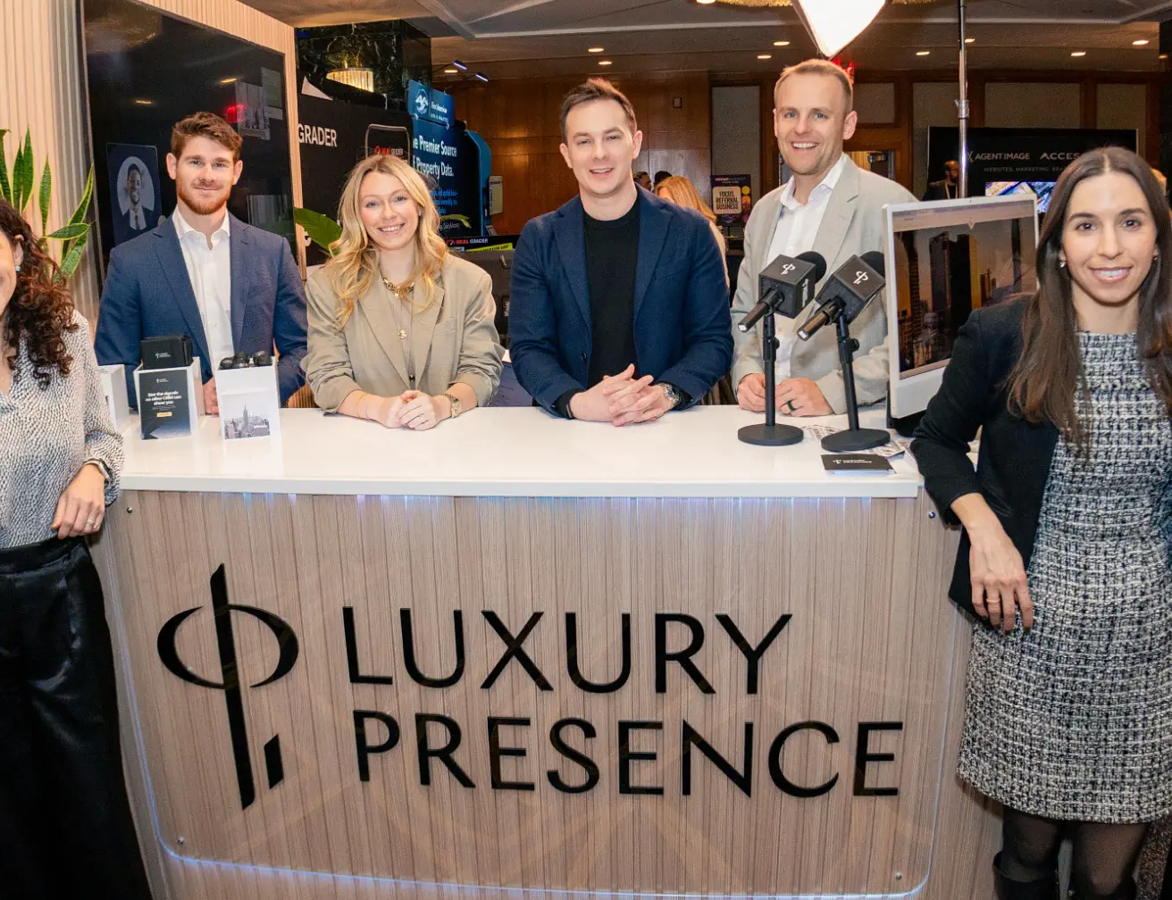 Five people smiling behind a desk with a 'Luxury Presence' sign at a professional event.