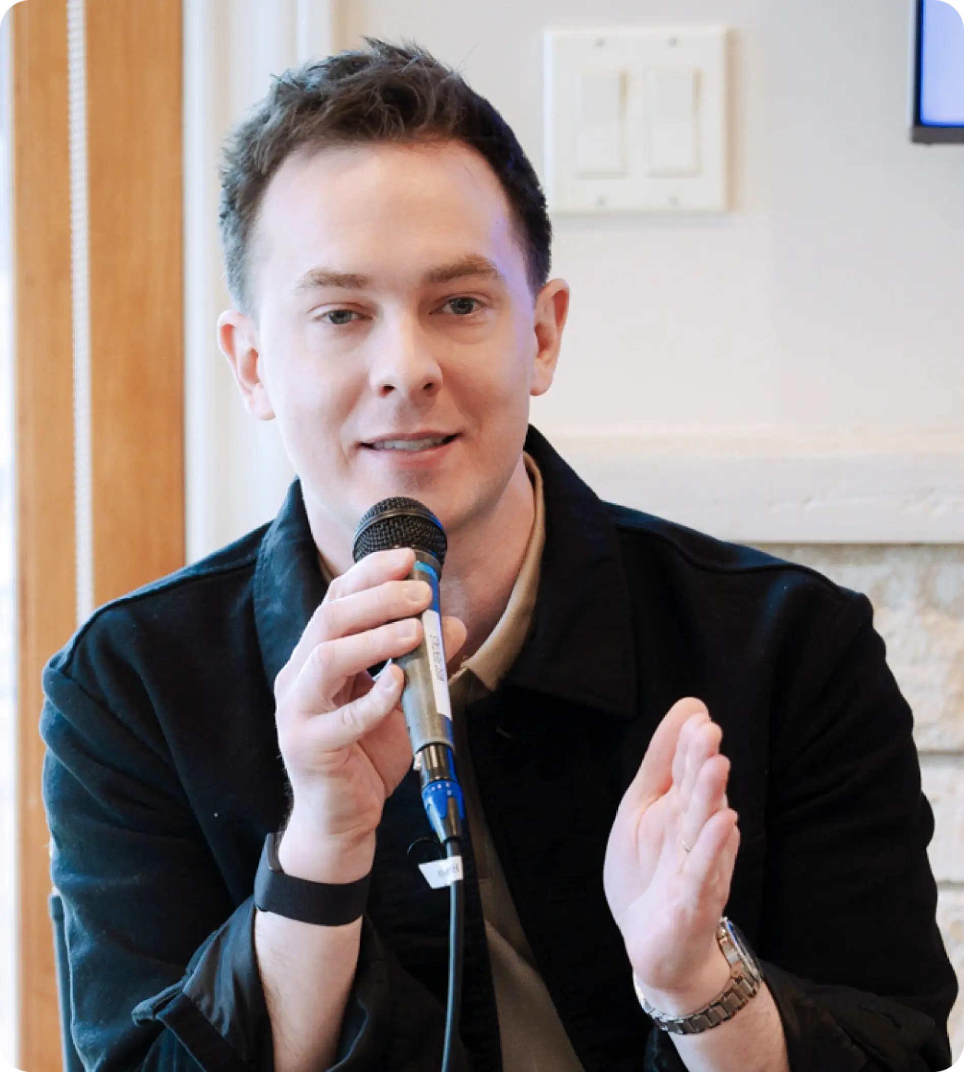 Kyle Scott holding a microphone speaking and gesturing with one hand while wearing a black jacket.
