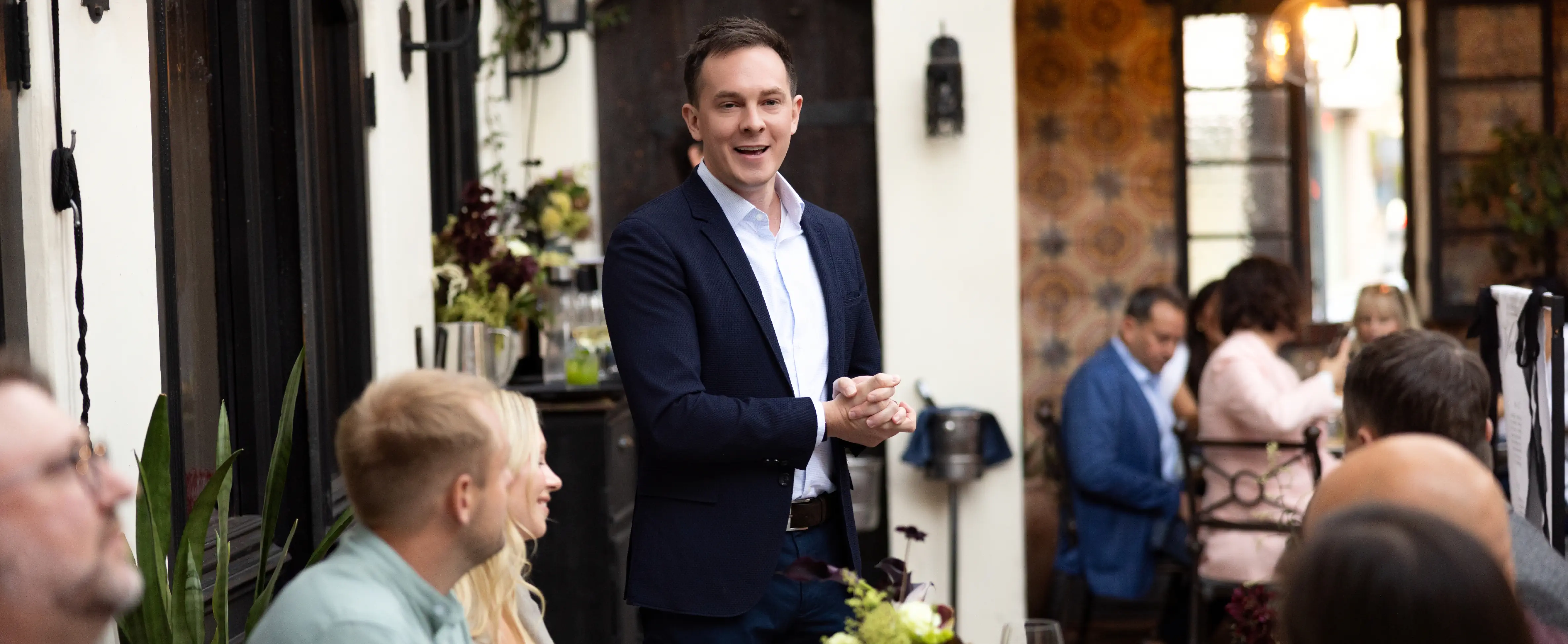 Kyle Scott in a dark blazer speaking to a group seated in a cozy indoor setting with plants and patterned walls.