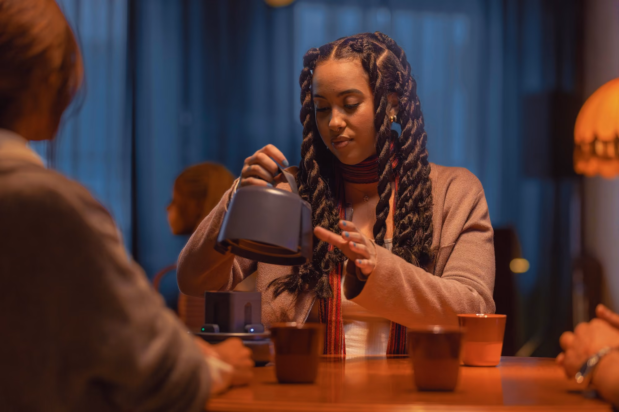 Young woman with braided hair pouring tea from a kettle at a table with three cups.