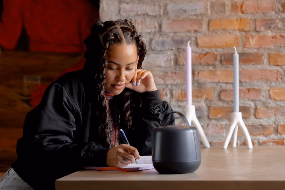 Young woman with braided hair writing in a notebook at a table with candles and a speaker.