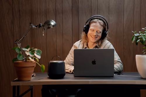 Person wearing headphones, smiling, working on a laptop at a desk with plants and a lamp.