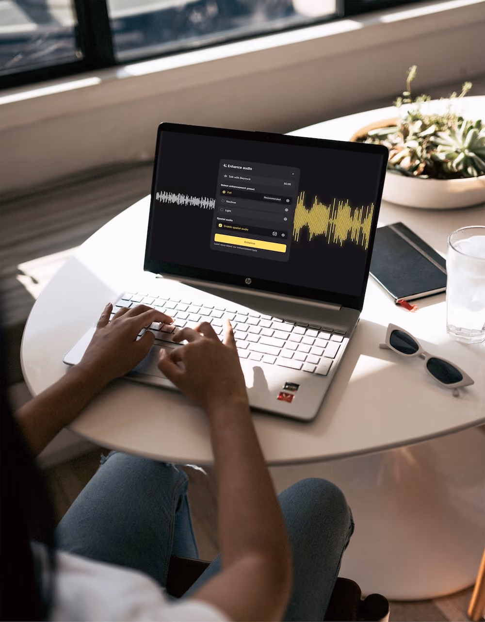 Person typing on laptop showing audio enhancement software with waveform and options on screen.