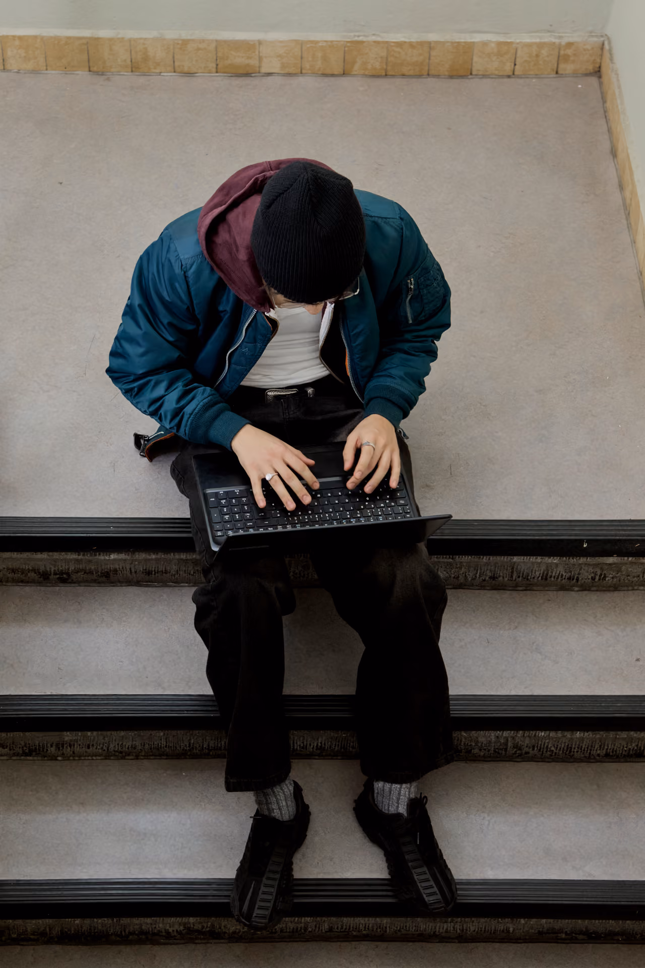 Person wearing a beanie and green jacket typing on a laptop while sitting on indoor stairs.