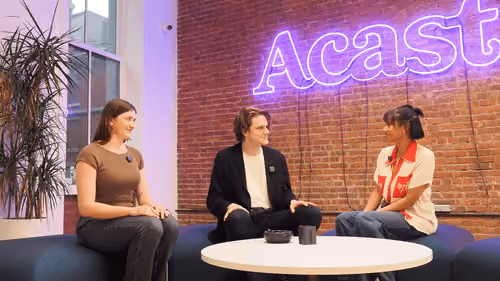 Three people sitting on a curved blue couch around a white table with a neon 'Acast' sign on brick wall.