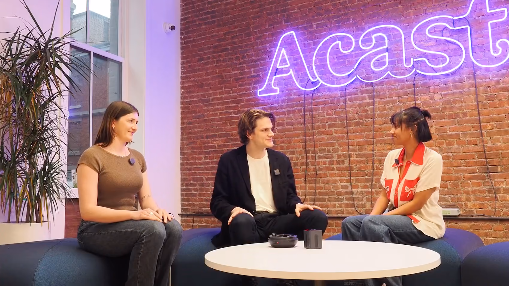 Three people sitting on a curved blue couch around a white table with a neon 'Acast' sign on brick wall.