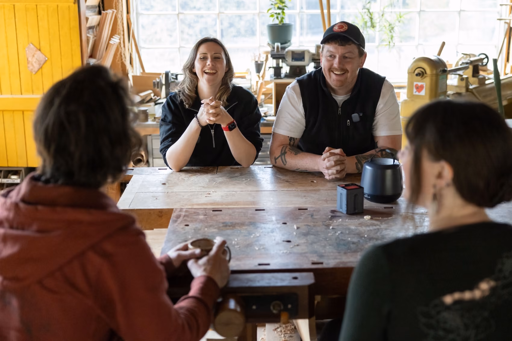 Two people smiling and talking across a workshop table to two others seen from behind.
