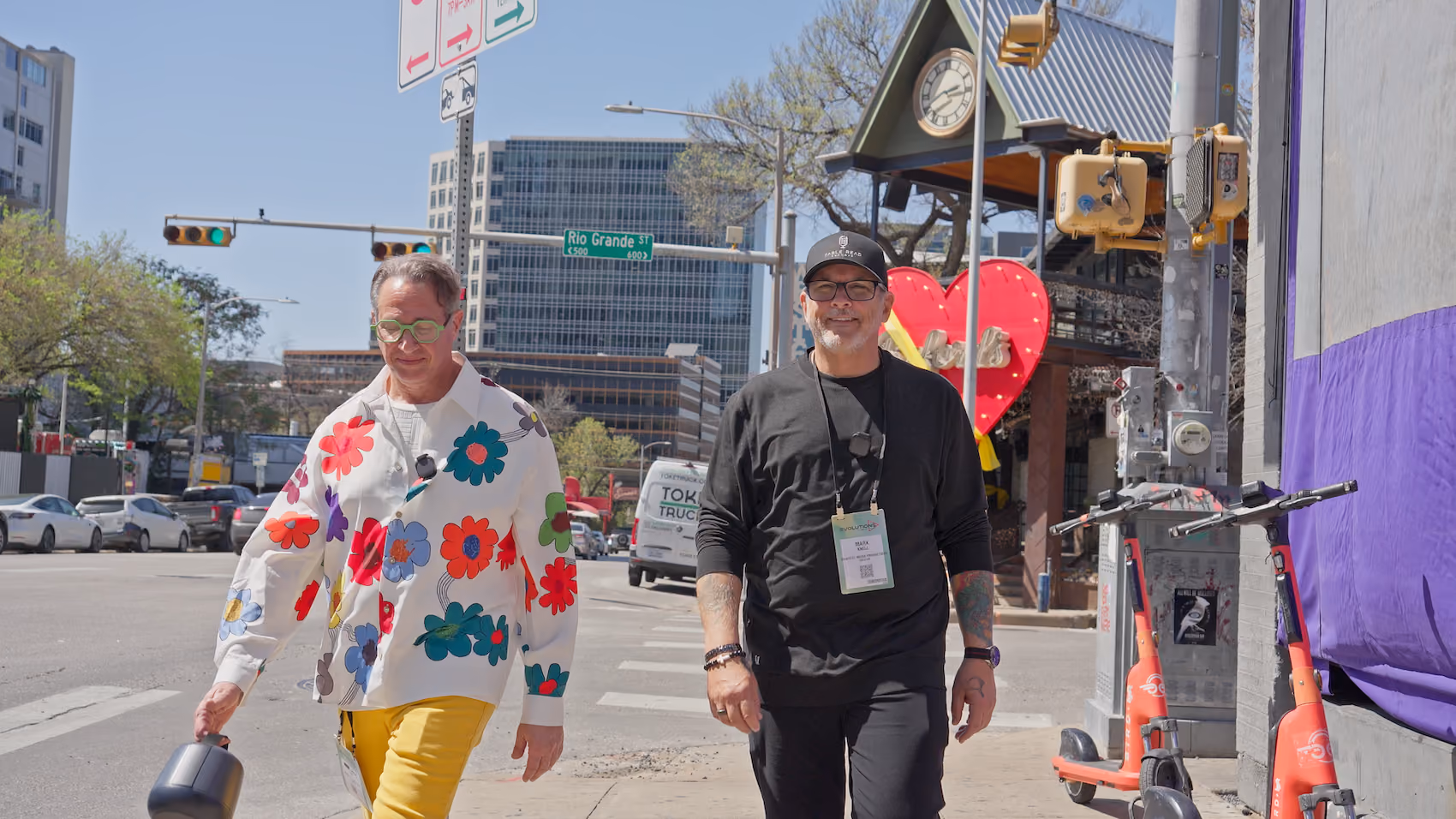 Two men walk on a city sidewalk; one wears a floral shirt and yellow pants, the other in black with tattoos.