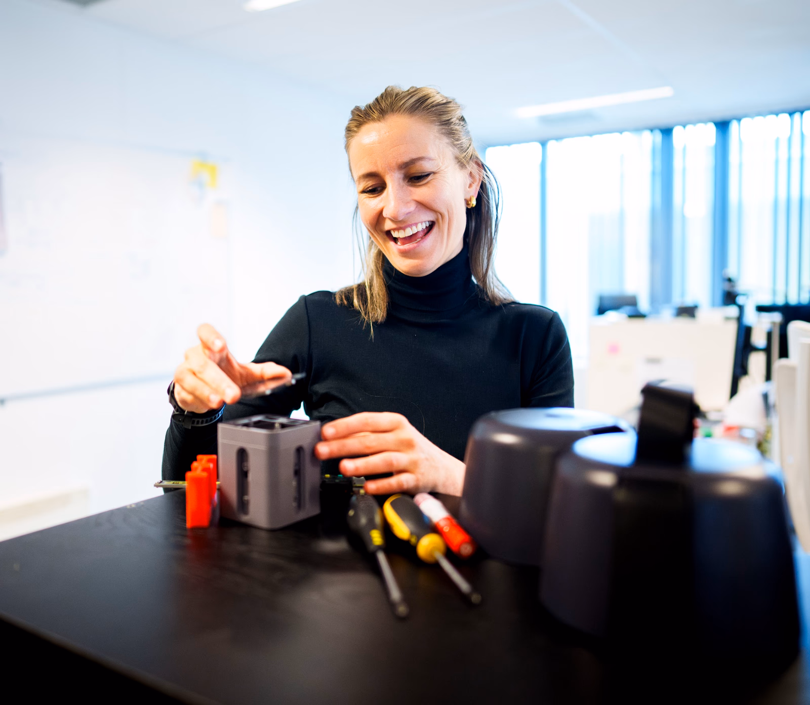 Smiling woman assembling a small mechanical device on a desk with tools in an office.