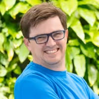 Smiling man with glasses and brown hair wearing a blue shirt, green leafy background.