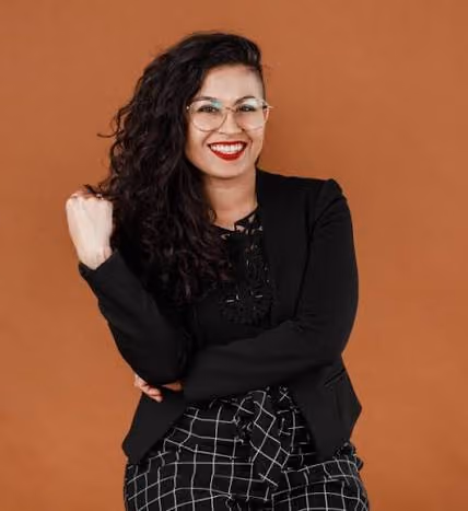 Smiling woman with curly hair and glasses wearing black blazer and checkered pants against brown background.