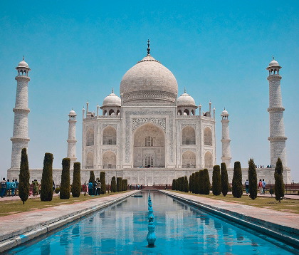 Front view of the Taj Mahal with reflecting pool and symmetrical minarets under clear blue sky.