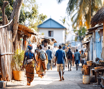 People walking down a sunlit village street lined with wooden huts and palm trees.