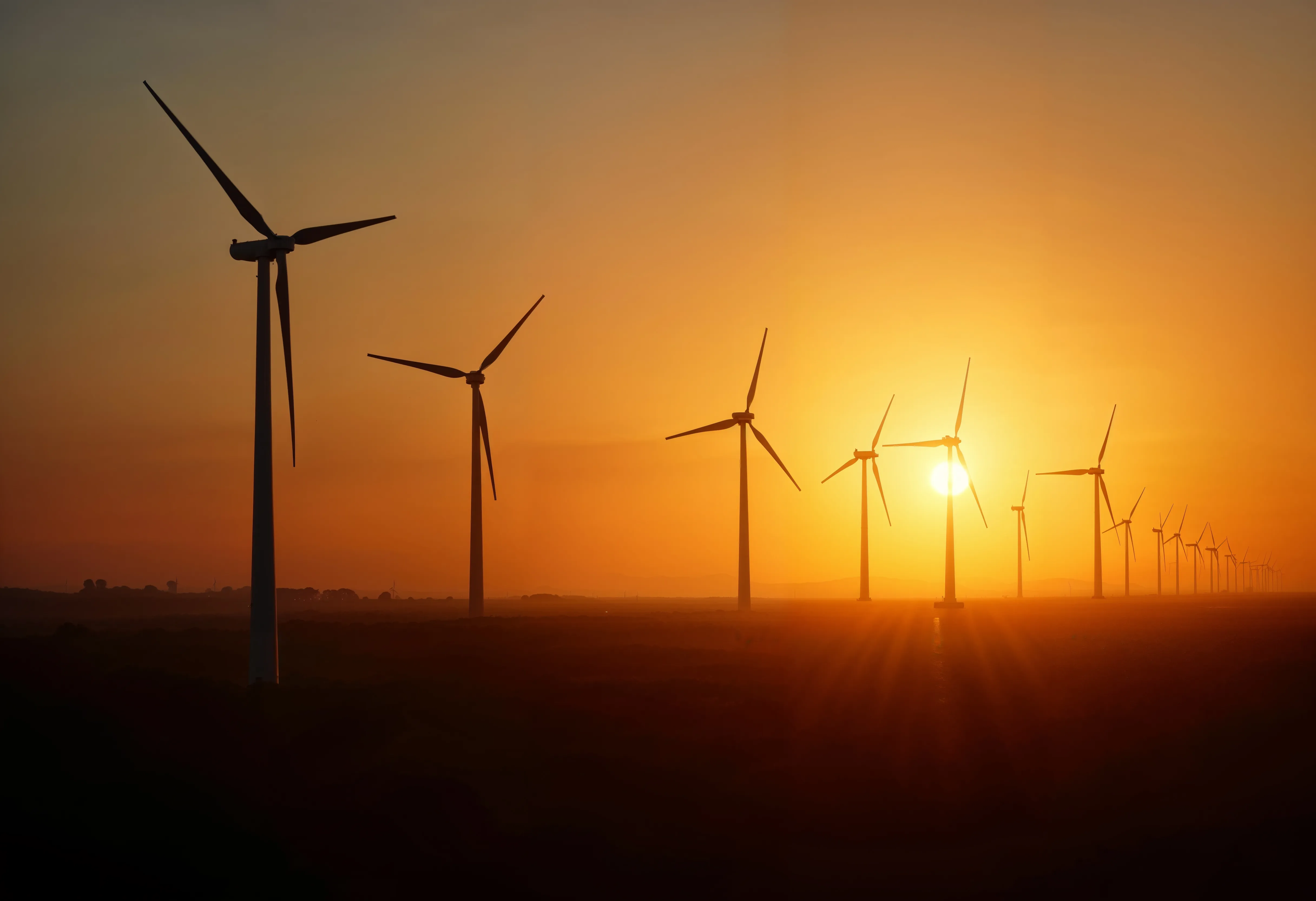 Line of wind turbines generating renewable energy at sunset with a bright orange sky.