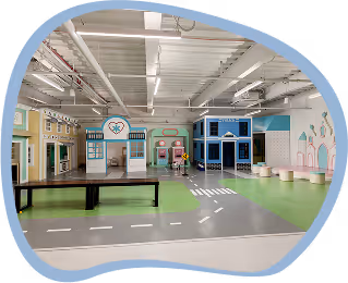 Indoor pretend play area with small storefronts, road markings, and seating for children.