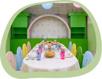 Colorful children's party room with a long table set for snacks and drinks, bright chairs and green shelves.