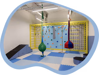 Indoor sensory gym with climbing wall, swings, ropes, and padded floor mats in blue and white.