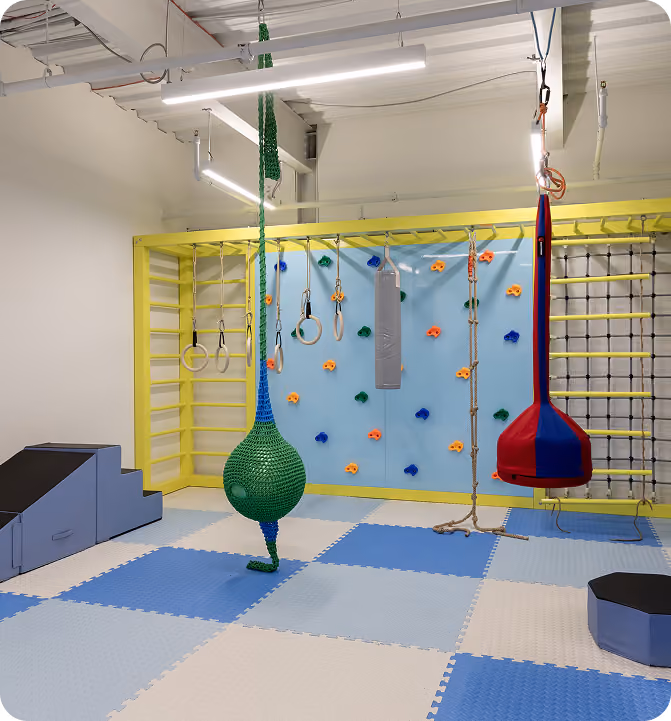 Indoor playroom with climbing wall, hanging rings, ropes, swings, padded floor, and play steps.