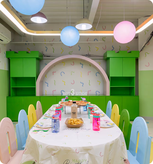 Colorful kids' birthday party room with a long table, pizza, fries, and bright balloons overhead.