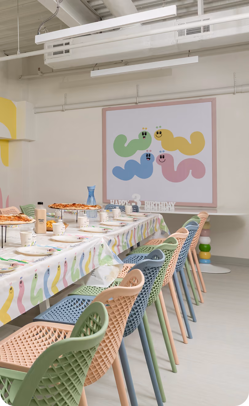 Long table set for a birthday party with colorful chairs and worm-themed decorations on the wall and table.
