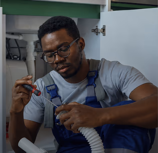 Man in glasses and overalls repairs plumbing under a sink using a screwdriver.
