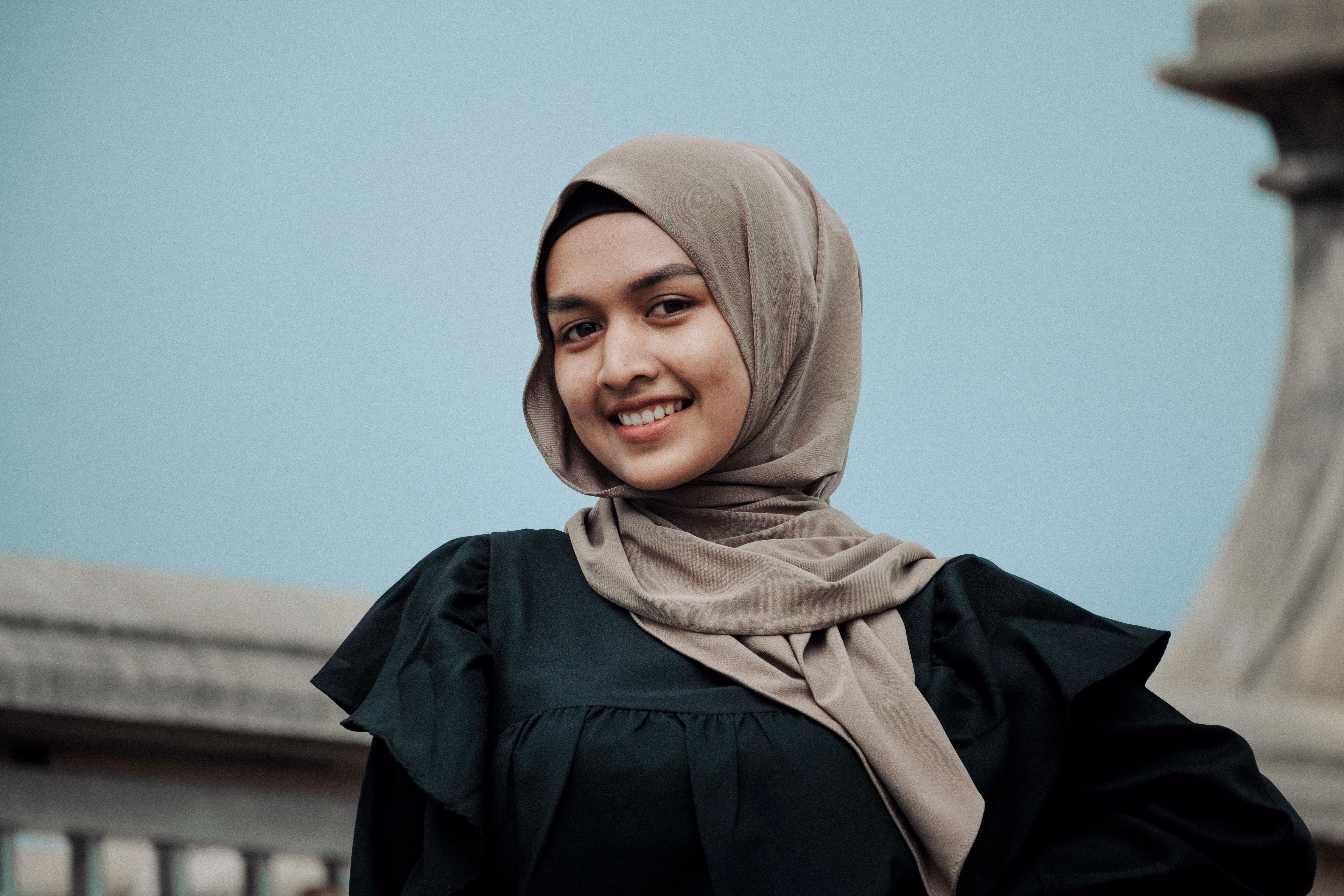 Young woman wearing a beige hijab and black dress smiling outdoors with a light blue background and stone railing.