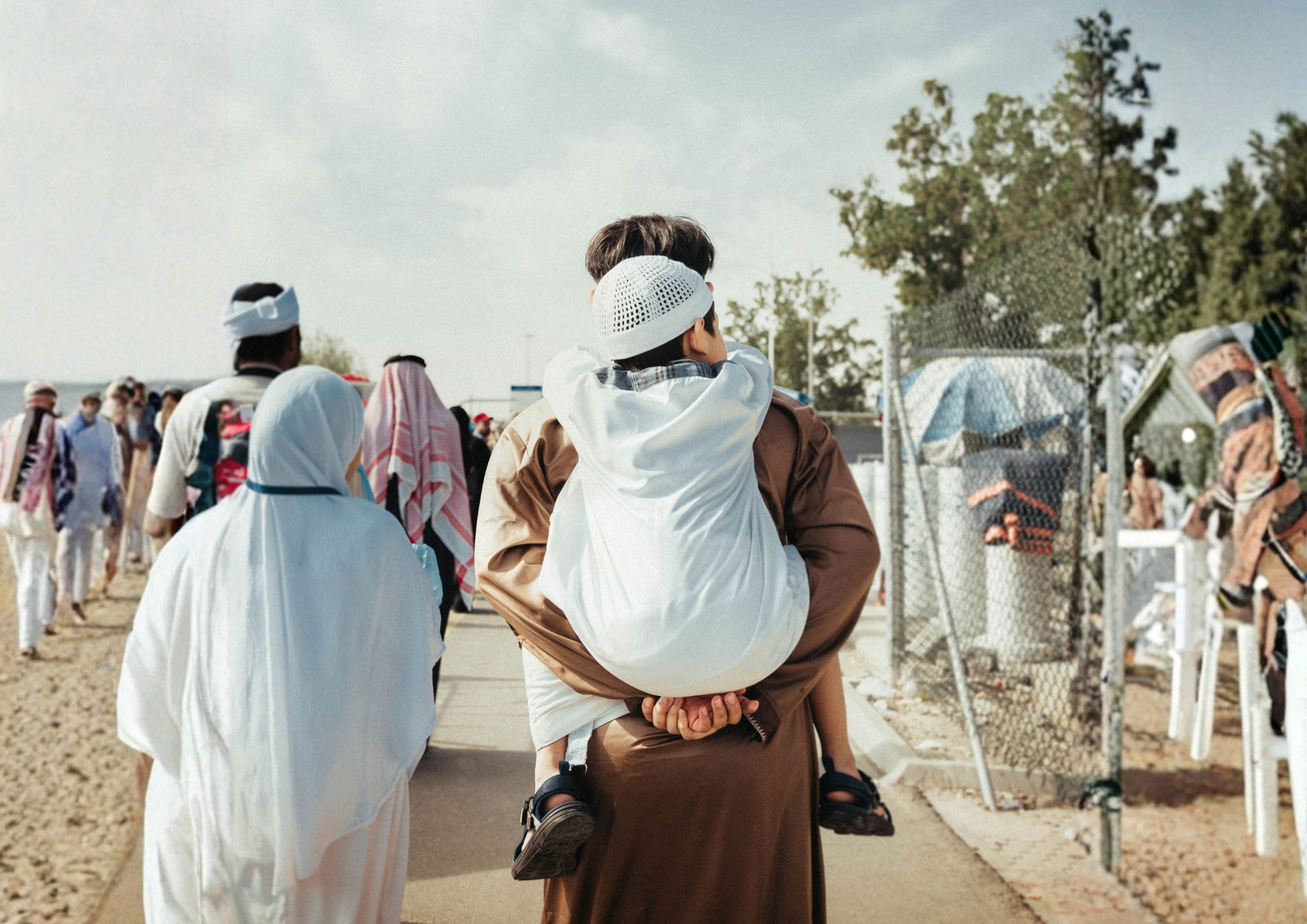 People walking on a paved path outdoors, with one person carrying a child on their back, both wearing traditional clothing including white garments and head coverings, and other individuals also dressed in traditional Middle Eastern attire.
