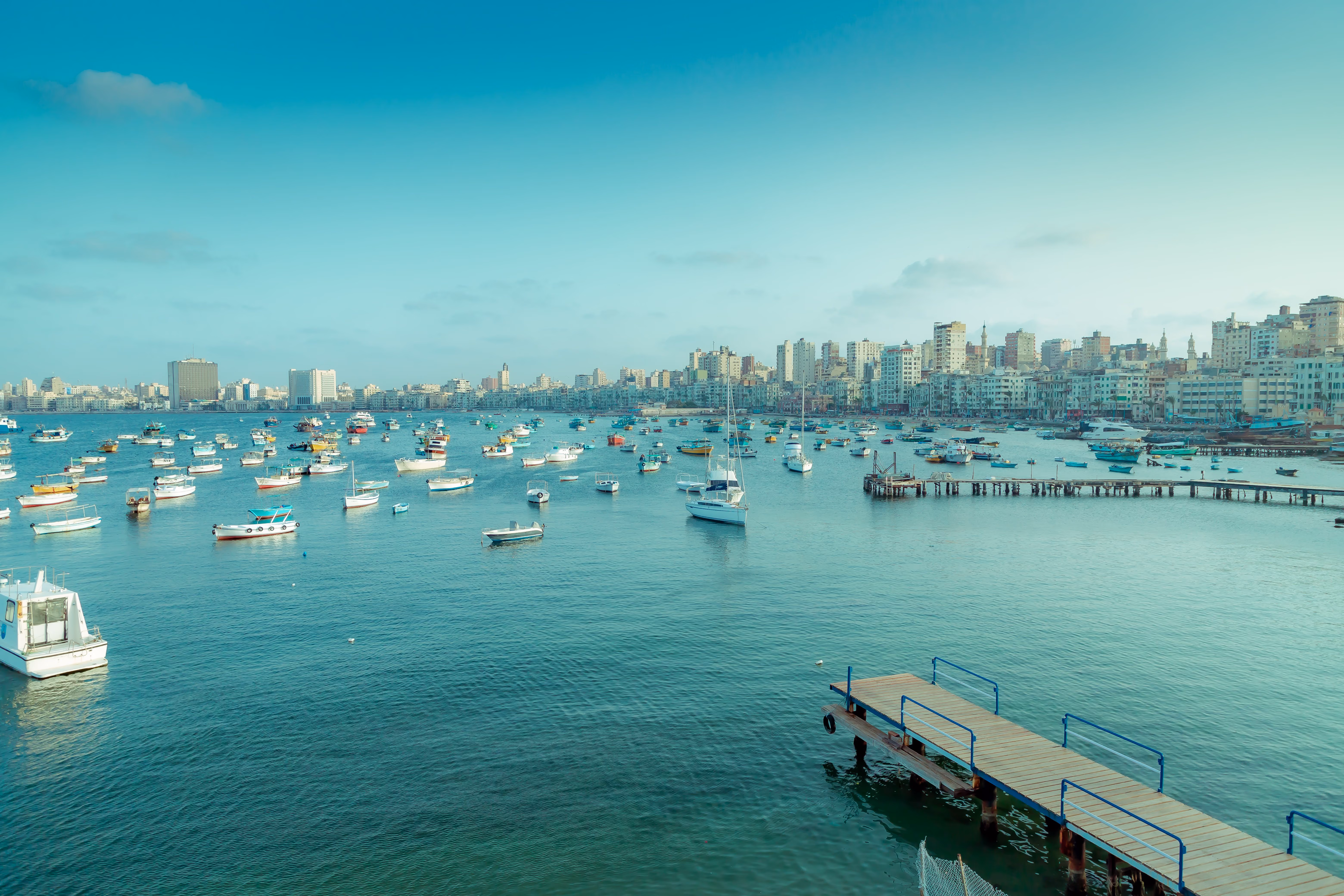 Calm coastal scene with many small boats anchored on turquoise water near a cityscape with numerous buildings under a clear sky, featuring wooden piers extending into the water.