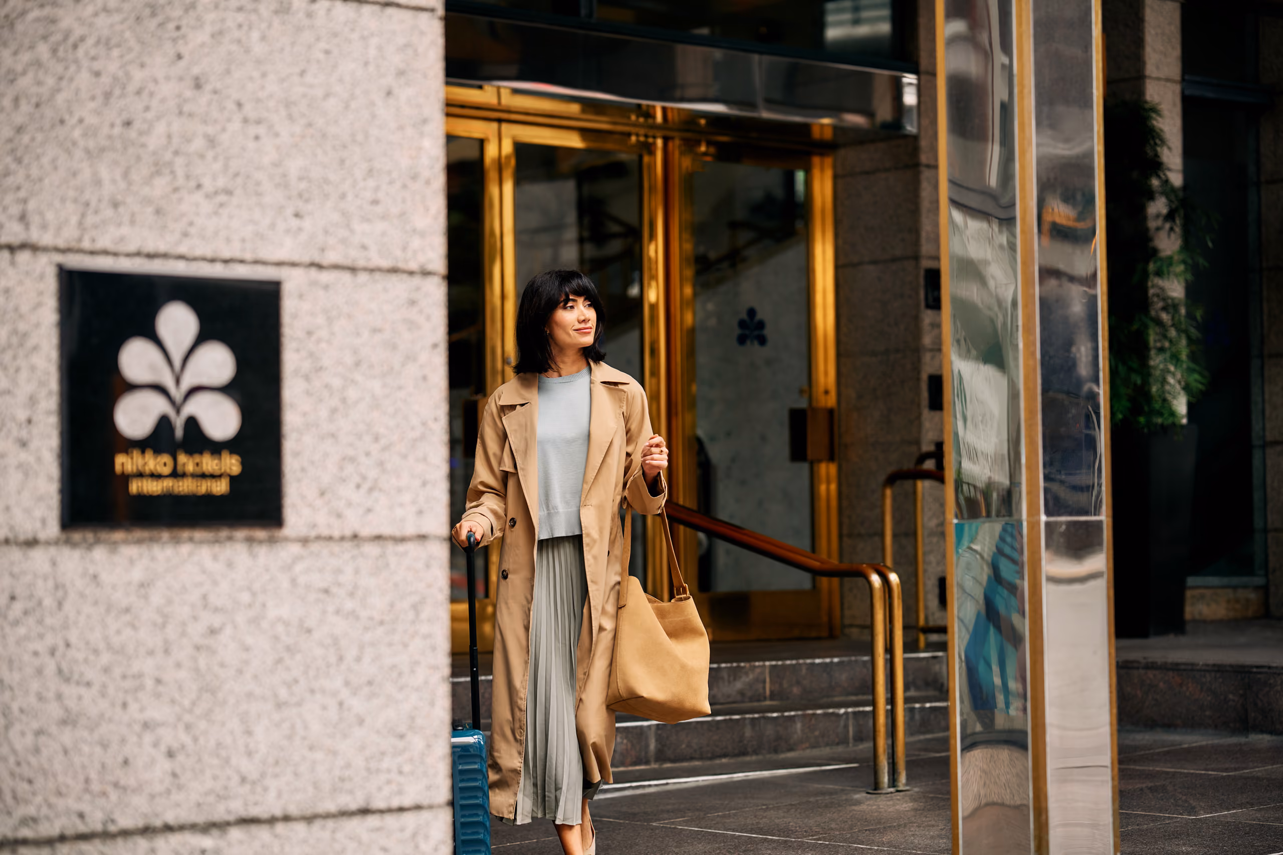 Guest outside hotel entrance with suitcase and tote bag.