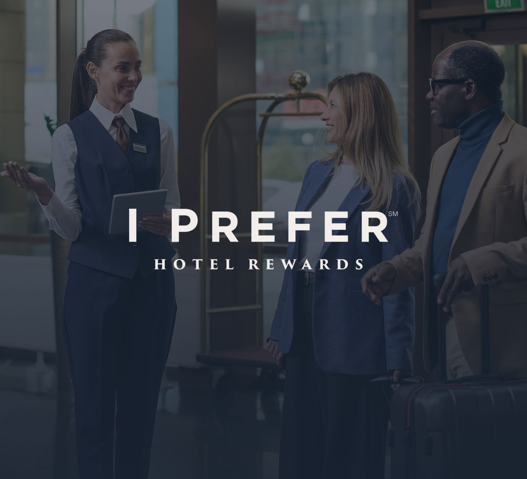 Hotel staff member greets two guests with luggage near a luggage cart at hotel entrance.